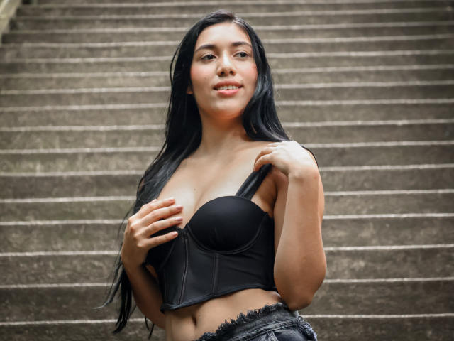 SaraRousse with long black hair, wearing a black strapless bra, holding one strap, showing her tits, and wearing frayed denim shorts, standing on a staircase.