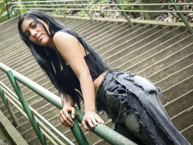 SaraRousse with long black hair, black crop top, and torn black pants, leaning on green rail, showing her ass, looking back with a slight smile.