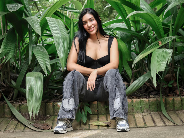 SaraRousse with black bra, large tits, grey ripped jeans, and silver sneakers, sitting on brick wall, smiling, long black hair, green leafy background.
