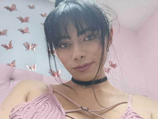 AnnaMooree, avec ses cheveux foncés, sa frange et son collier, porte un haut rose en crochet, laissant entrevoir de petites seins, avec du rouge à lèvres rose et un sourire discret, dans une pièce rose décorée de papillons..