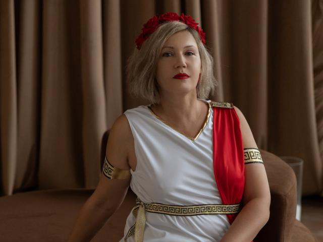 A photo of NinaStarle with shoulder-length, wavy, gray hair and a red flower headpiece. She's wearing a white toga-style dress with a red sash, gold armlet, and Greek key-patterned belt. She has red lipstick and a neutral expression. She's seated on a brown chair against brown curtains. The image has a classical, ancient Greek theme with simple, warm colors.