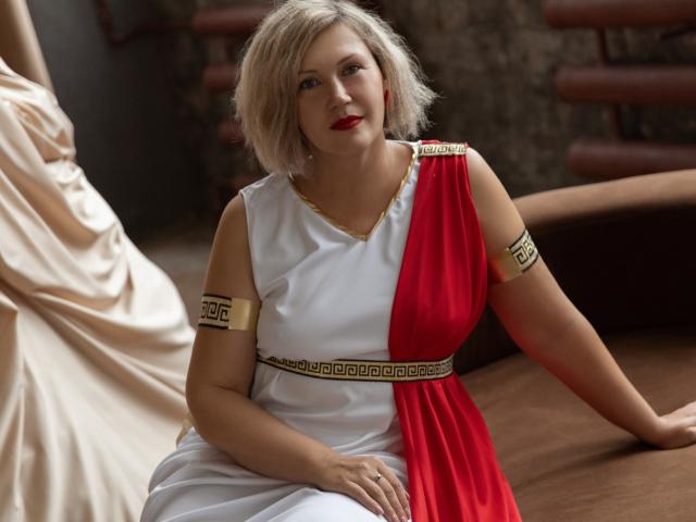 NinaStarle, a fair-skinned woman with short blonde hair, wears a Greek-style dress with a red and white split. The dress has a gold armband with a geometric pattern and a gold belt with a similar design. She sits on a brown surface, her red lipstick contrasting with her white and red dress. The background is blurred, focusing attention on NinaStarle. The texture of the dress is smooth, with a slight sheen. The composition is balanced, highlighting her relaxed, confident pose.