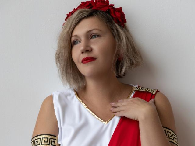 NinaStarle met kort blond haar, rode rozen in haar haar, een rood-witte jurk met gouden details, rode lippenstift, hand op de borst, licht omhoog kijkend, witte achtergrond..