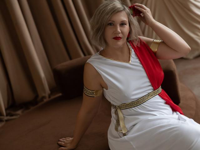 NinaStarle, a blonde woman with a shoulder-length wavy hairstyle, sits on a brown couch. She wears a white toga-style dress with a red sash on her right shoulder, gold arm bands with Greek key patterns, and a gold belt with a patterned sash. She has red lipstick and is holding a red flower in her right hand. The photo is a close-up shot, focusing on her upper body and face. The lighting is soft, highlighting her fair skin and the textures of the fabric. The background features brown, draped curtains.