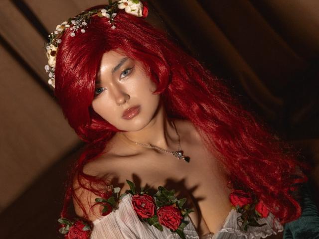 AnneTLuis with red wavy hair, flower crown, wearing a white off-shoulder dress with red roses, showing her tits, pale skin, and a necklace with a small black pendant.