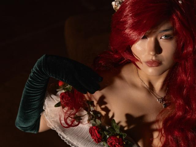 AnneTLuis with red hair, green velvet gloves, white lace dress, red roses, and a necklace, looking up with a serious expression, soft light, dark background.