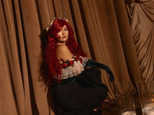 AnneTLuis is a cosplay character with long, wavy red hair, wearing a floral headpiece. She's dressed in a strapless white top with red roses, green velvet gloves, and black pants. She's crouching with a serious expression, set against brown, draped curtains. Glass cups are on the floor to her right. The lighting is warm, casting soft shadows. The overall composition is balanced, with a focus on AnneTLuis's detailed costume.