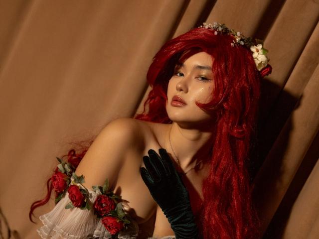 AnneTLuis with red wavy hair, flower crown, green gloves, and white dress with red roses, exposing her tits, looking sexy with a slight smile, against a brown curtain.