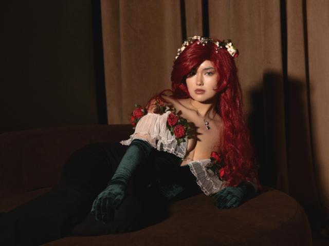 AnneTLuis with red hair, wearing a dark Victorian dress, green gloves, flower crown, and rose decorations, lounging on a brown sofa, showing her tits and a necklace.