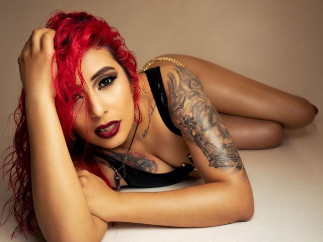 MegaanGrey with bright red hair, tattoos on her right arm, and dark makeup, lying on her side in black lingerie, looking at the camera with red lipstick.