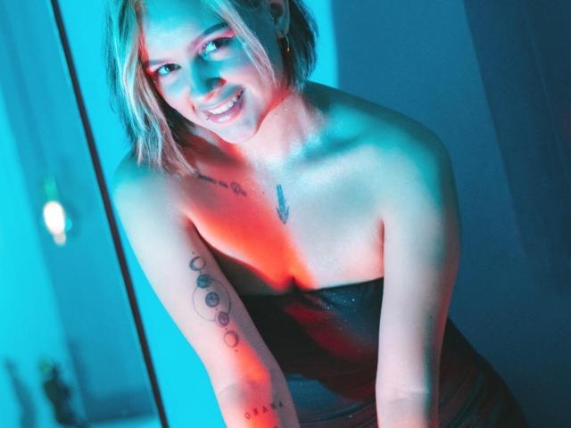 IsabellaMontier tiene el cabello corto y rubio, y está sonriendo a la cámara. Lleva un vestido oscuro y brillante, sin tirantes. Su piel es clara, y tiene tatuajes en el brazo derecho, incluyendo una línea vertical de círculos y una pequeña pluma en el pecho. La imagen está iluminada con luces azules y rojas, creando un efecto dramático y colorido. Su expresión es amigable y acogedora. La fotografía utiliza un alto contraste y un enfoque nítido..