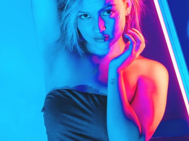 IsabellaMontier met een stralend blauw en roze neonlicht op haar gezicht, haar hand die haar wang aanraakt, gekleed in een glanzend zwart topje zonder bandjes, blond haar, een subtiele glimlach, tatoeages op haar vingers..