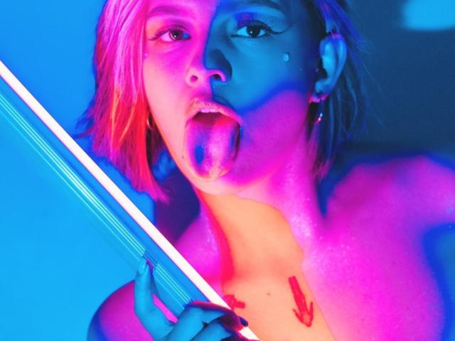IsabellaMontier met roze en blauwe neonverlichting, tong uit, een neonstok vasthoudend, kleine tatoeages op de borst, kort roze kapsel, oor piercings, naar de camera kijkend, topless, glanzende huid..