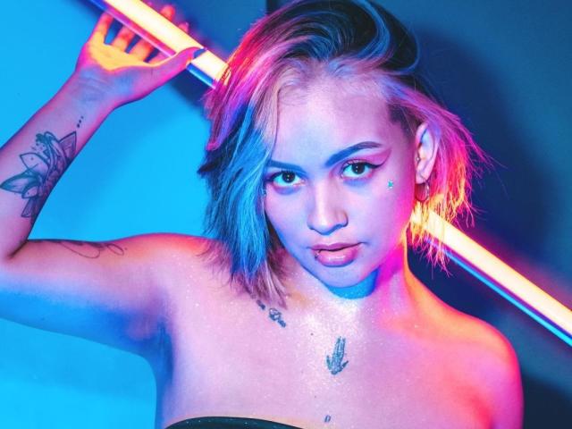 IsabellaMontier ha i capelli di lunghezza media, parzialmente biondi e parzialmente tinti di blu. Tiene in mano con la mano destra una barra verticale illuminata al neon, mostrando un tatuaggio con un motivo geometrico sulla parte superiore del braccio. Ha un piccolo tatuaggio sotto l'occhio destro e un altro tatuaggio sul petto. La sua pelle è leggermente lucida e indossa un top senza spalline. La luce al neon blu e rosa proietta ombre colorate sul suo viso e sul suo corpo. La sua espressione è neutra, con le labbra leggermente dischiuse. La composizione generale utilizza colori vivaci e contrasti netti..