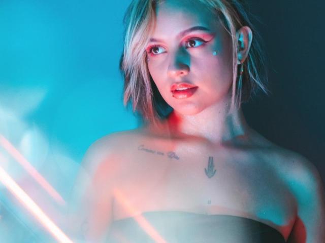 IsabellaMontier met blond haar, stralende blauwe en roze neonlichten, die borsten en tatoeages op de borst laat zien, kijkt in de camera met rode oogmake-up en een subtiele glimlach..