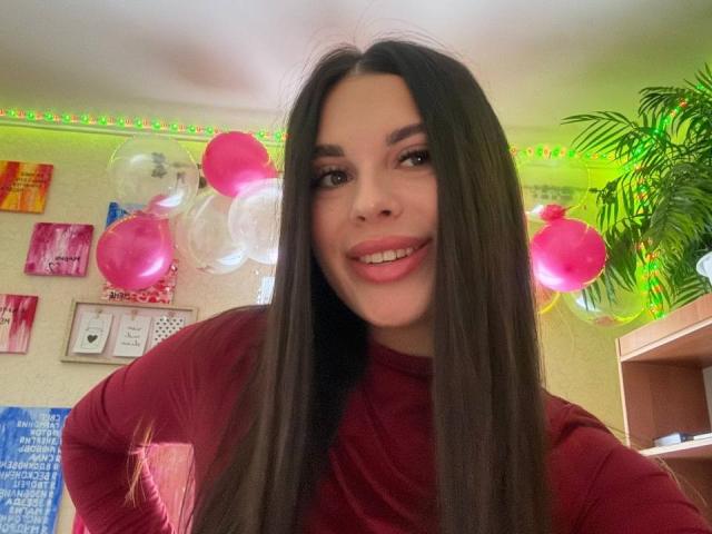 BlackFatal, con cabello castaño largo, camisa roja, sonriendo a la cámara, globos rosas y blancos en el fondo, luces LED verdes y una planta en maceta a la derecha..