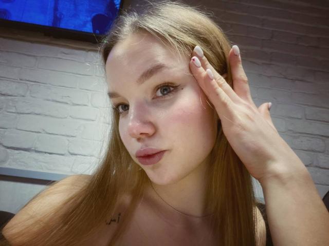 StellaLights with blonde hair, blue eyes, and pink lips, touching her forehead with a hand. She's wearing a necklace, and her nipples are slightly visible under her top.