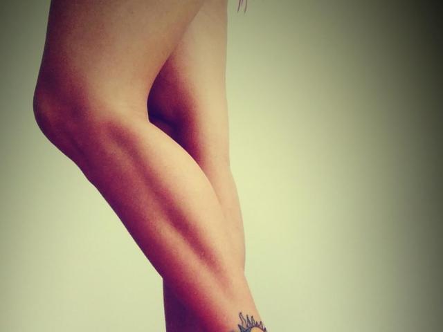 RosannaBlue's leg with a tattoo on the ankle, showing smooth skin, a slight bend, and a subtle shadow against a neutral background.