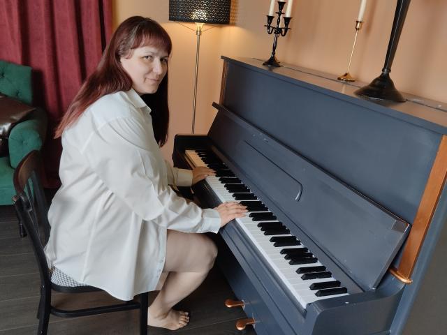 AliceInks sits on a black chair at a dark blue upright piano. She's a middle-aged woman with long red hair, wearing a white button-up shirt, and white underwear is visible. She's barefoot, with her right hand on the piano keys and left on the piano's front. The piano has black and white keys, and a wooden top. Behind her is a green upholstered chair and a red curtain. The lighting includes a black lamp and white candles. The floor is dark wood.