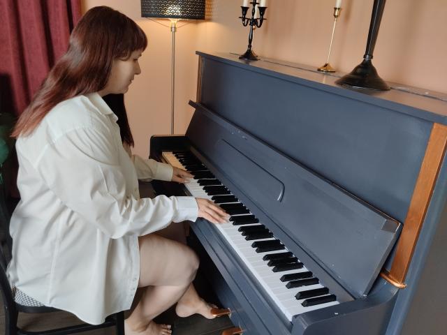 AliceInks with brown hair, white shirt, and bare legs, playing a blue upright piano, her hands on the keys, her pussy not visible, in a room with candles.