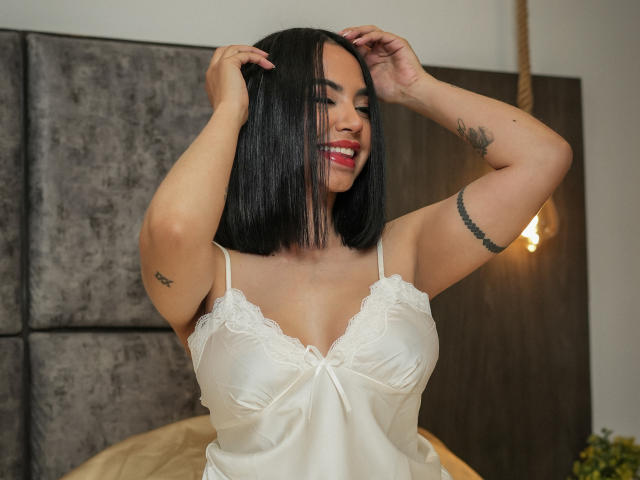 SweetstLady with black bob, smiling, hands in hair, wearing white lace-trimmed satin nightie, showing small tattoos, big tits, and red lipstick. Bedroom setting, warm light.