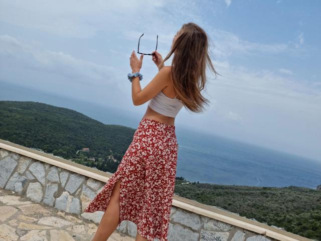Leilaaa-hot with long brown hair, white crop top, and red floral skirt, holding glasses, standing on stone terrace, looking at ocean, wearing blue scrunchie.