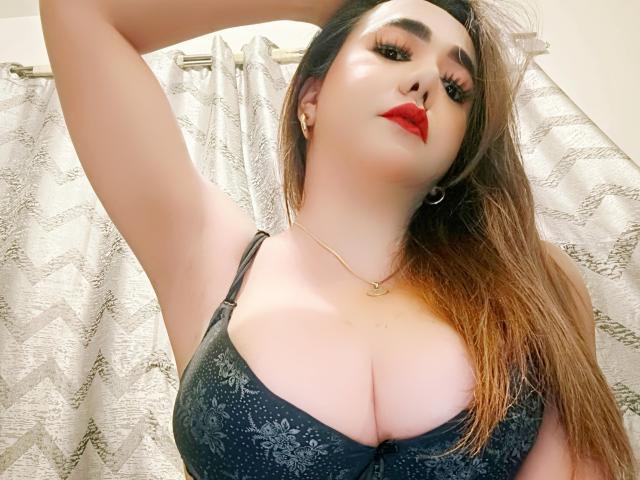 FabulousNina69 with big tits in a black lace bra, red lipstick, and long brown hair, arm raised, looking down, wearing hoop earrings and a gold necklace.