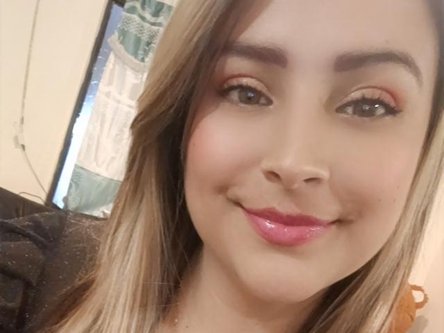 Marcee69 has light brown hair, pink lipstick, and a cute smile. Her brown eyes are slightly upturned with orange eyeshadow. She's wearing a black top.
