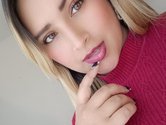 Marcee69 with blonde hair, pink lipstick, and red sweater, touching her pink lips with a finger, showing dark nail polish, and looking at the camera with brown eyes.