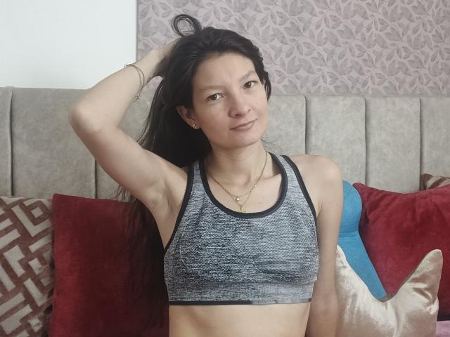 LilyHottie69 with long black hair, wearing a grey sports bra, hand in hair, small tits, light skin, subtle smile, gold necklace, sitting on a bed with red and blue pillows.