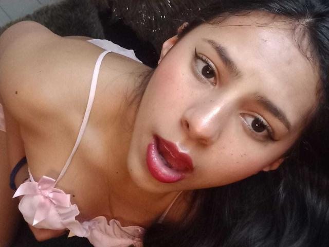 SoffiRousee has shiny skin, dark eyes, and red lips, wearing a pink bra with a bow, looking up with her mouth slightly open, her black hair spread out.