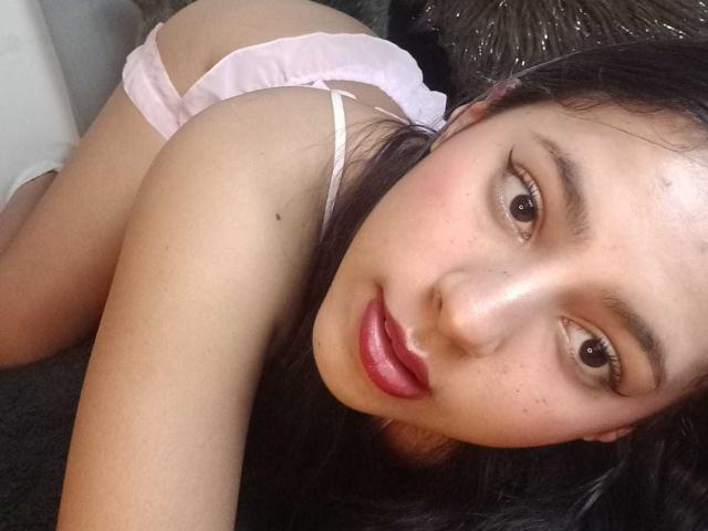 SoffiRousee is on her knees, wearing pink panties, with her ass up and face looking at the camera. She has dark hair, red lips, and a cute freckled face.