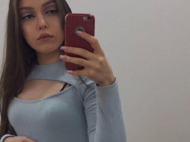 Miss-Merciless-hot with long brown hair, wearing a light blue top with cut-out chest, holding a red phone, showing her small tits, looking serious, pink nails, white background.