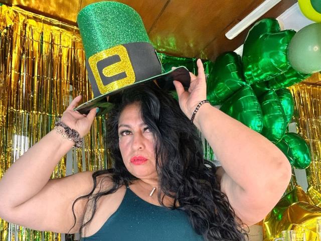Keyla69Hot, con un sombrero verde brillante de duende, cabello ondulado negro, lápiz labial rojo, blusa verde, sosteniendo el sombrero, globos dorados y verdes, serpentinas doradas, mirando a la cámara, ligeramente rechoncha..