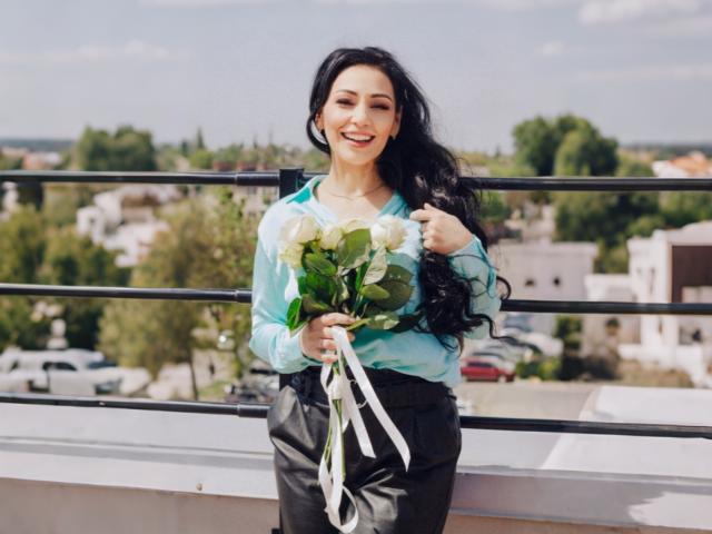 LiluGrants, con lunghi capelli neri, sorridente, indossa una camicia color turchese e pantaloni neri, tiene in mano rose bianche con foglie verdi e nastri bianchi, ed è in piedi su un balcone..