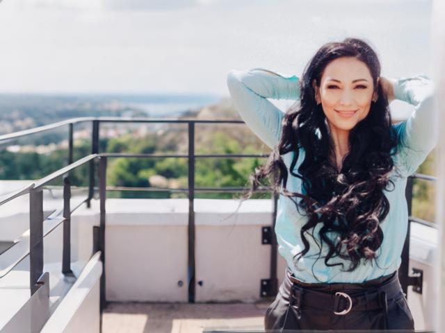 LiluGrants with long, wavy black hair, smiling, wearing a light blue shirt and black pants, standing with hands in hair, on a sunlit balcony.