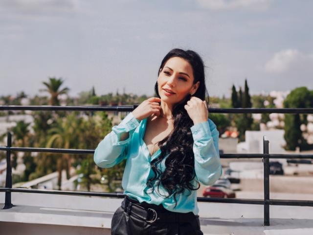 LiluGrants with long black hair, wearing a light blue shirt, black pants, and a belt, standing on a balcony, hands near her face, smiling.