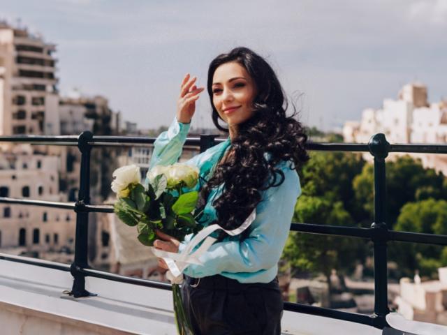 LiluGrants with long curly black hair, wearing a light blue shirt, holding a bouquet of white and green flowers, standing on a balcony with black railings.