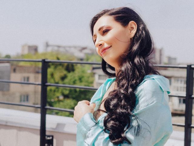 LiluGrants with long black braids, wearing a shiny blue top, holding her chest, smiling, standing on a balcony with black railings, in bright daylight.