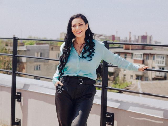 LiluGrants, with long black curly hair, wearing a light blue shirt and black pants with a crescent moon belt, standing and smiling against a black metal railing.