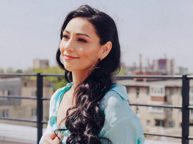 LiluGrants with long, wavy black hair, light skin, and a cute smile, wearing a blue top, standing on a rooftop, looking to the side, showing her tits.