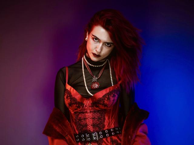 CosmoAngy met rood haar, donkere lipstick, gekleed in een rode kant korset, zwarte mesh mouwen, een parelketting en een riem met studs, voor een achtergrond met een blauw-paarse gradiënt..