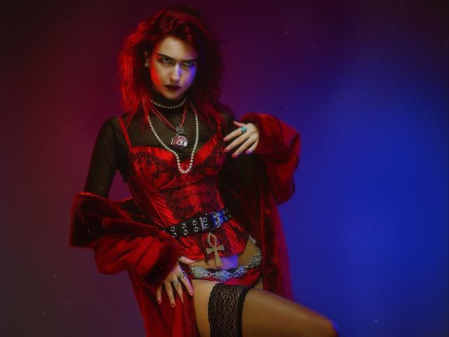 CosmoAngy, con cabello rojo, vistiendo un corpiño de encaje rojo, mangas largas negras, un abrigo de piel roja, medias de encaje negras hasta el muslo, un collar de perlas y un cinturón, luciendo seductora, bajo luces azules y rojas..