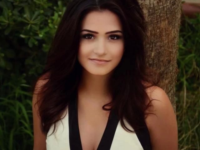 LiaMia-hot has dark, wavy hair, brown eyes, and a subtle smile. She's wearing a sleeveless, black-and-white top, standing in front of a tree.