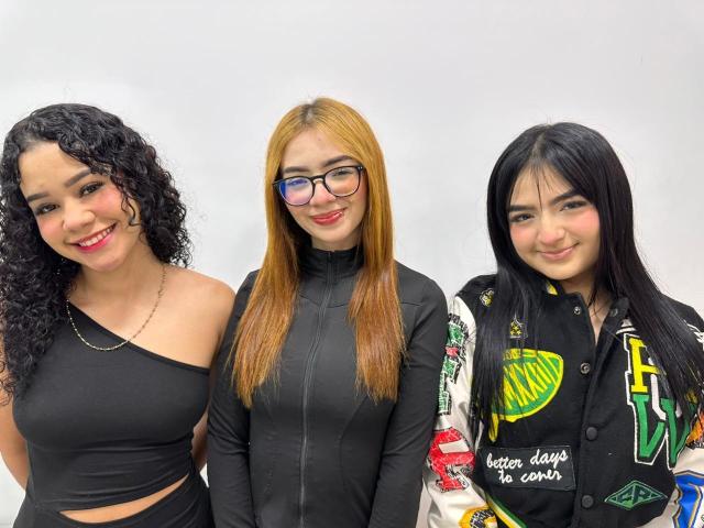 Three GlamorousHotties standing, black outfits, curly brunette, straight blonde with glasses, straight black-haired, cute smiles, black jacket with colorful patches, white background, casual, friendly, attractive.