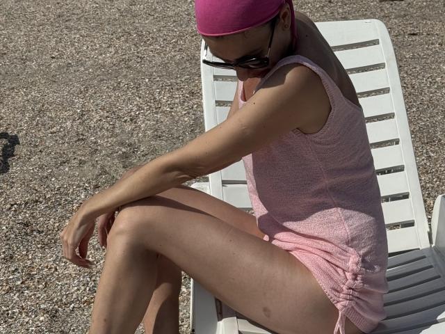 Nalini with pink cap, sunglasses, pink tank top, and shorts, sitting on white chair, legs crossed, arms resting, pebbled ground, sunlight, tan skin, relaxed, casual.