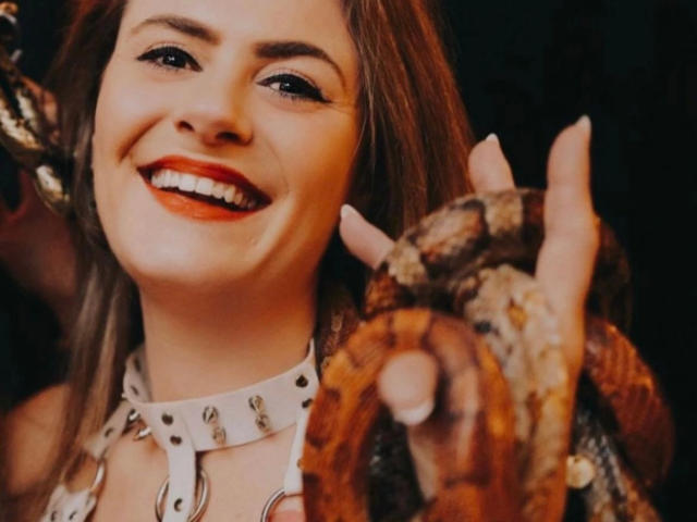 BadAnna-hot with red lipstick, smiling, holding a snake's head in her right hand, wearing a white studded collar and snake-skin gloves.