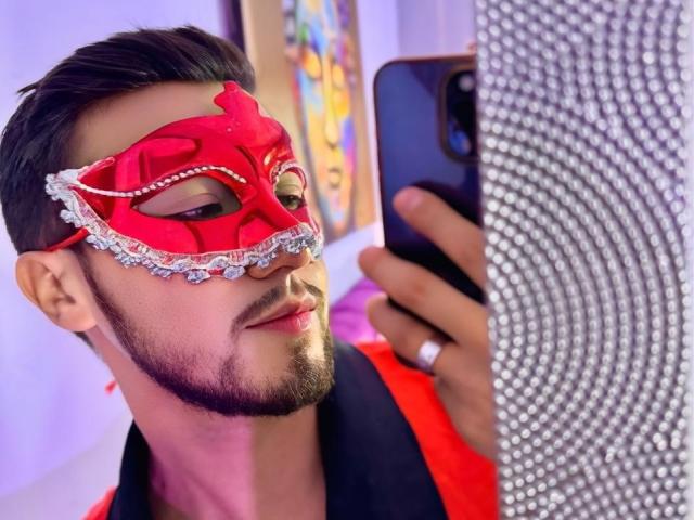 DaltonWhite portant un masque de bal rouge, des ornements en diamant et une barbe, prenant un selfie avec un téléphone noir, vêtu d'une chemise rouge et portant une alliance à sa main droite..