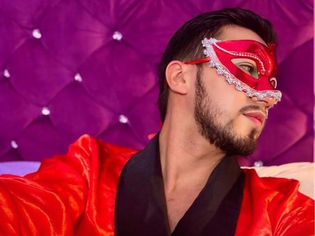 DaltonWhite, con una maschera rossa bordata di pizzo, indossa una veste rossa lucida, ha una barba corta e capelli scuri, e guarda di lato con un leggero sorriso..