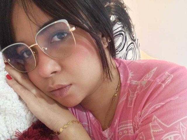 AliceSmity with puffy pink cheeks, wearing gold-framed glasses, pink shirt, and gold bracelet, resting her head on a white pillow, looking slightly pouty. Her nails are painted red.